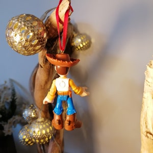 May include: A small plastic figurine of Woody from Toy Story hanging from a red ribbon. Woody is wearing his signature brown hat, yellow shirt, blue jeans, and brown boots. He is facing away from the camera.