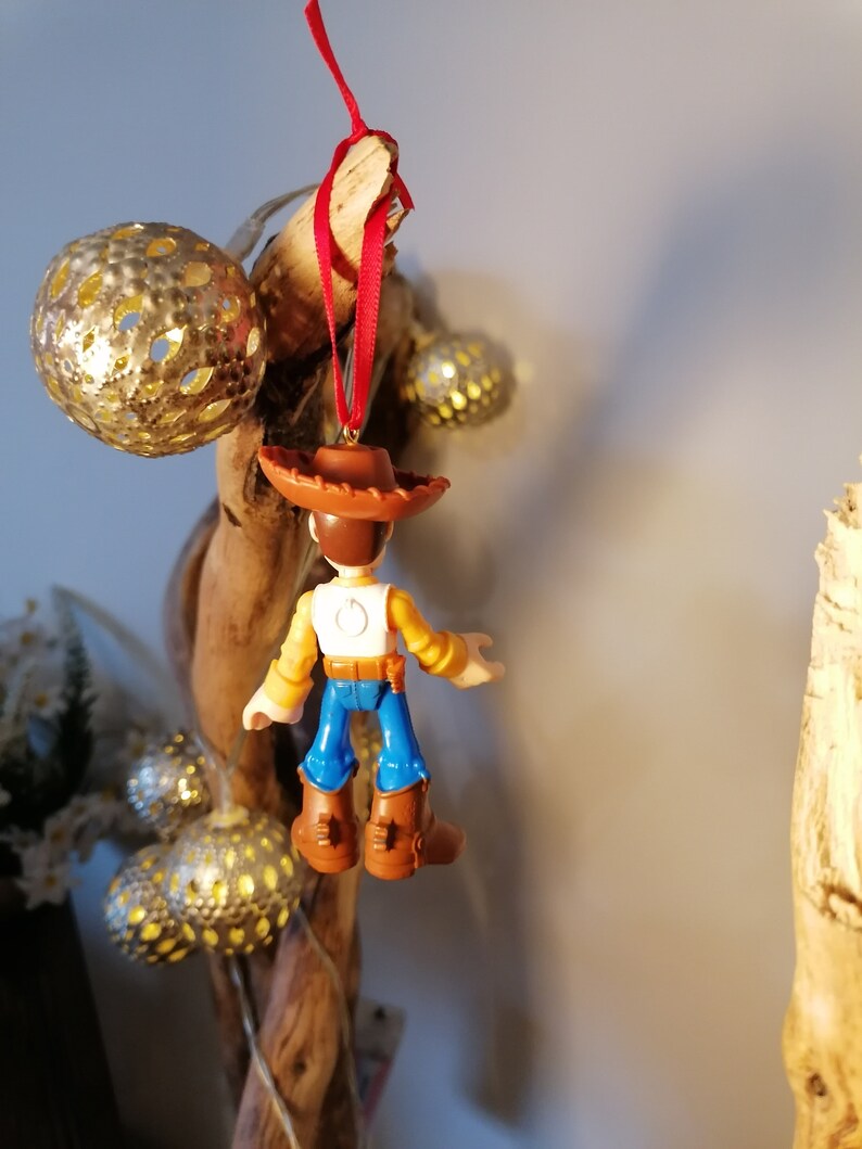 May include: A small plastic figurine of Woody from Toy Story hanging from a red ribbon. Woody is wearing his signature brown hat, yellow shirt, blue jeans, and brown boots. He is facing away from the camera.