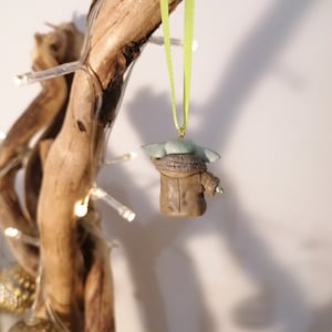 Pu&ograve; includere: Una statuetta verde e marrone di un personaggio del franchise di Star Wars, appesa a un nastro verde su un ramo di legno galleggiante.