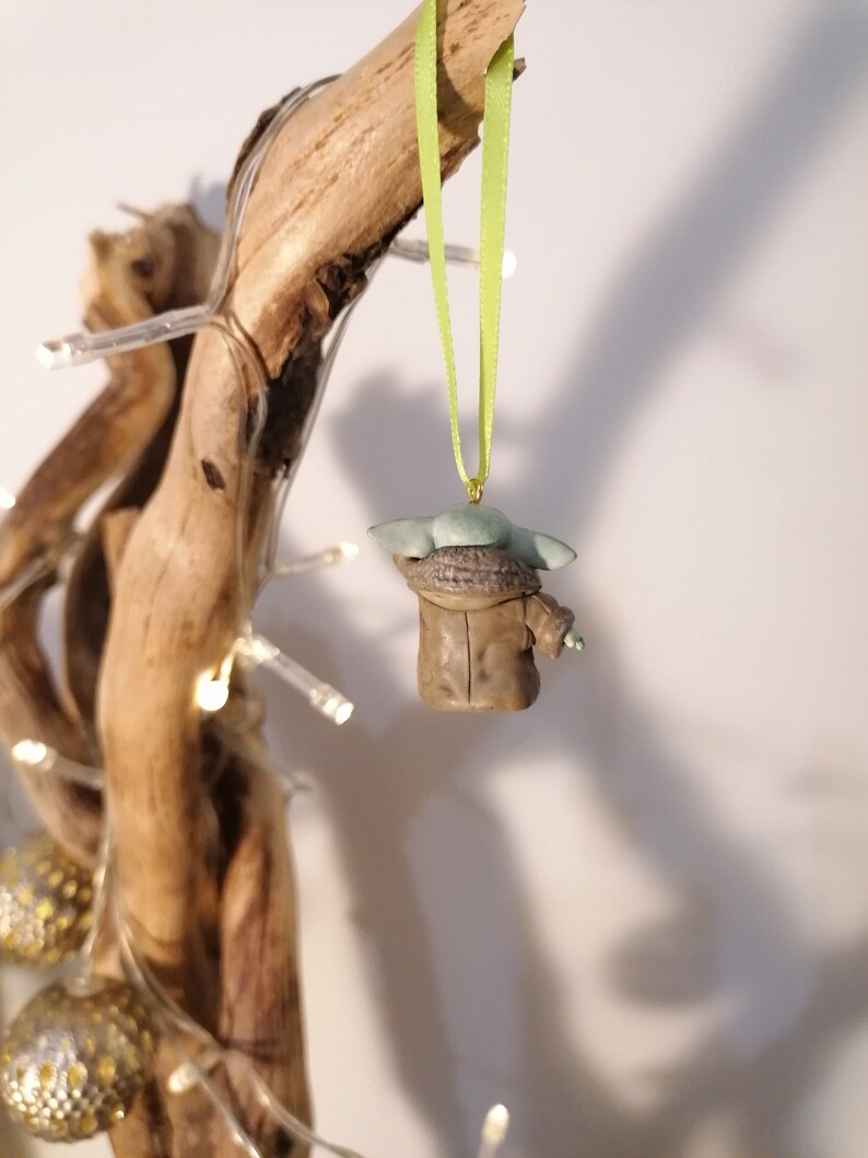 Pu&ograve; includere: Una statuetta verde e marrone di un personaggio del franchise di Star Wars, appesa a un nastro verde su un ramo di legno galleggiante.