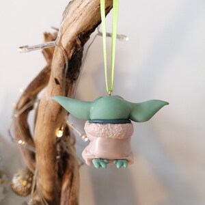 May include: A green and brown Christmas ornament shaped like a character from the Star Wars franchise. The ornament is hanging from a green ribbon.