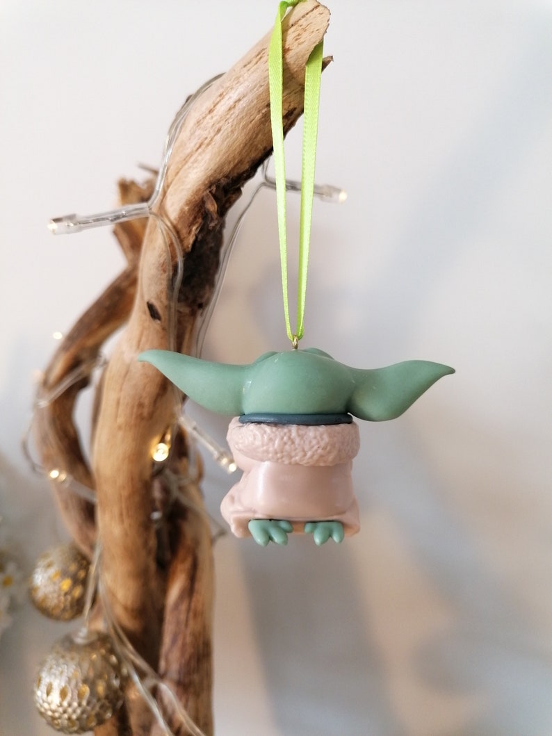May include: A green and brown Christmas ornament shaped like a character from the Star Wars franchise. The ornament is hanging from a green ribbon.