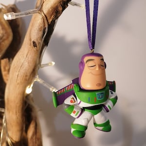 K&ouml;nnte beinhalten: Ein gr&uuml;ner und lila Buzz Lightyear Weihnachtsbaumschmuck mit einem lila Band. Der Schmuck ist eine stilisierte Version des Charakters aus den Toy Story Filmen.