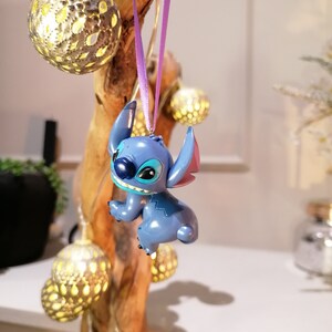 Pode incluir: Adorno de Natal azul Stitch pendurado em uma fita roxa em um galho de madeira com luzes decorativas douradas.