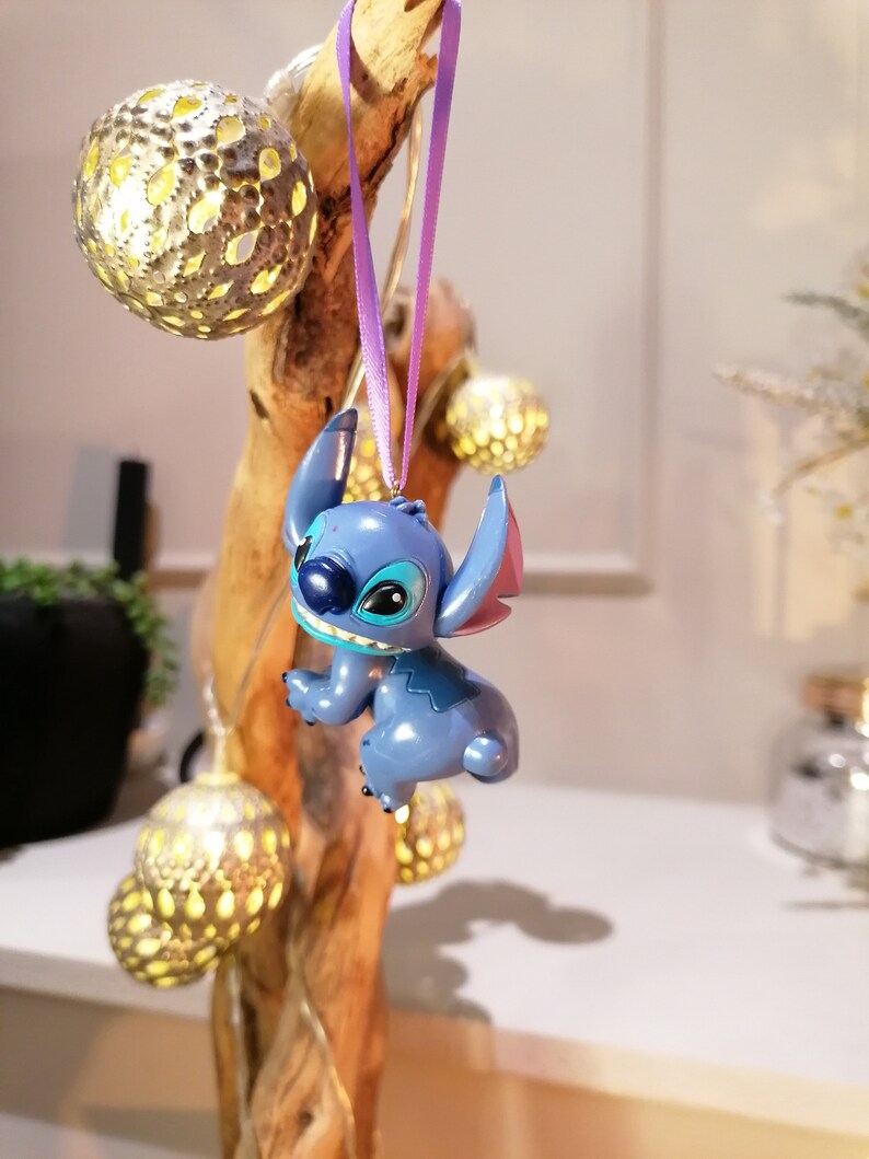 Pode incluir: Adorno de Natal azul Stitch pendurado em uma fita roxa em um galho de madeira com luzes decorativas douradas.