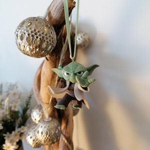 May include: A green Yoda ornament hanging from a branch with a green ribbon. The ornament is made of plastic and has a detailed design.