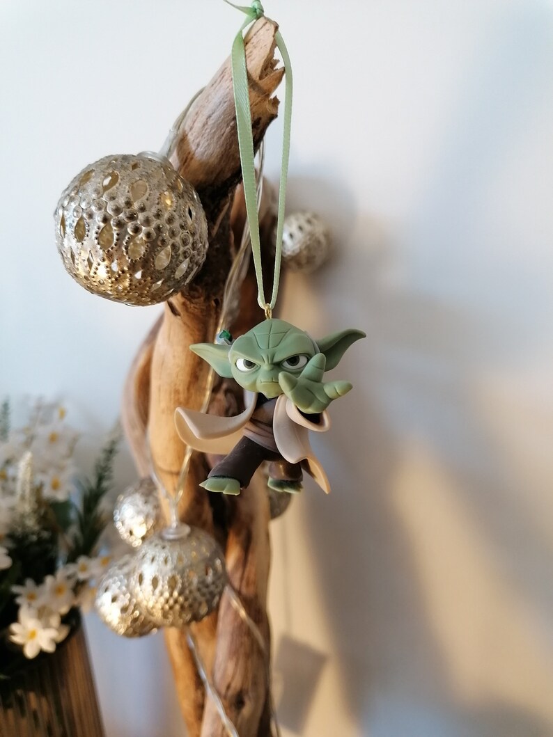 May include: A green Yoda ornament hanging from a branch with a green ribbon. The ornament is made of plastic and has a detailed design.