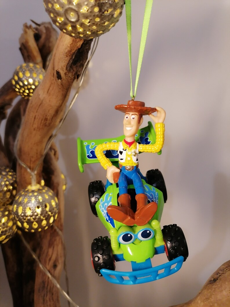 May include: A green toy race car with a blue bumper and a blue and green design.  Woody from Toy Story is sitting in the driver's seat, wearing his signature cowboy hat and vest.  The car is hanging from a green ribbon.
