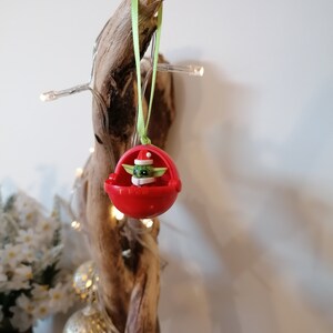 Pu&ograve; includere: Un ornamento natalizio rosso con un piccolo personaggio verde che indossa un cappello da Babbo Natale. L'ornamento &egrave; appeso a un nastro verde su un pezzo di legno alla deriva.