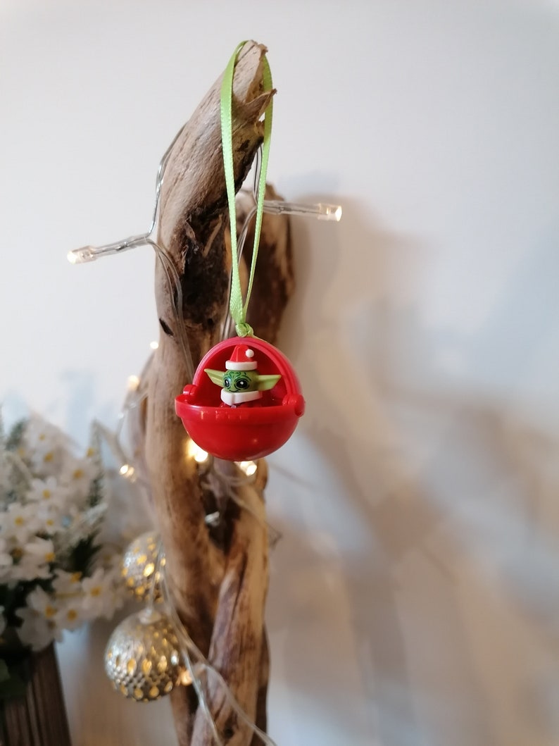 Pu&ograve; includere: Un ornamento natalizio rosso con un piccolo personaggio verde che indossa un cappello da Babbo Natale. L'ornamento &egrave; appeso a un nastro verde su un pezzo di legno alla deriva.