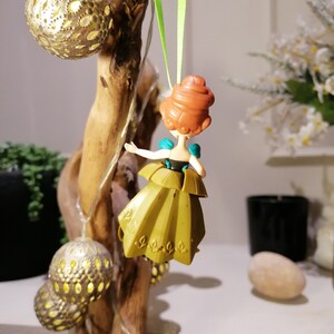 May include: A green and gold dress ornament featuring a cartoon character with red hair. The ornament is hanging from a green ribbon.
