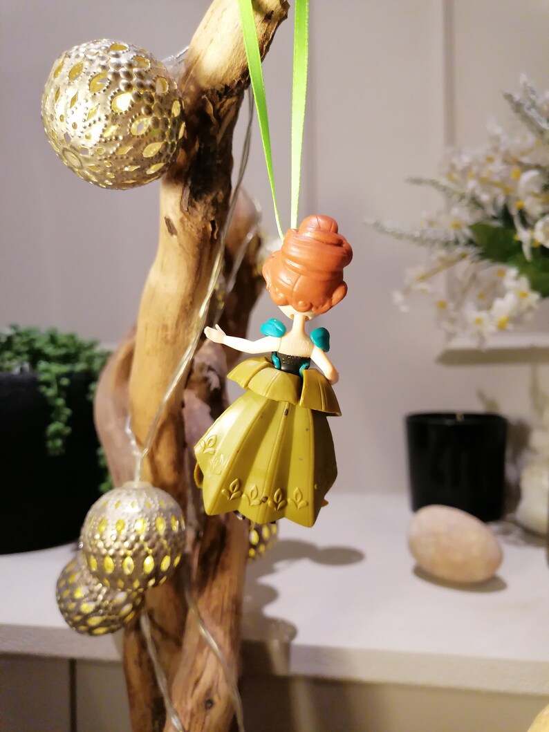 May include: A green and gold dress ornament featuring a cartoon character with red hair. The ornament is hanging from a green ribbon.