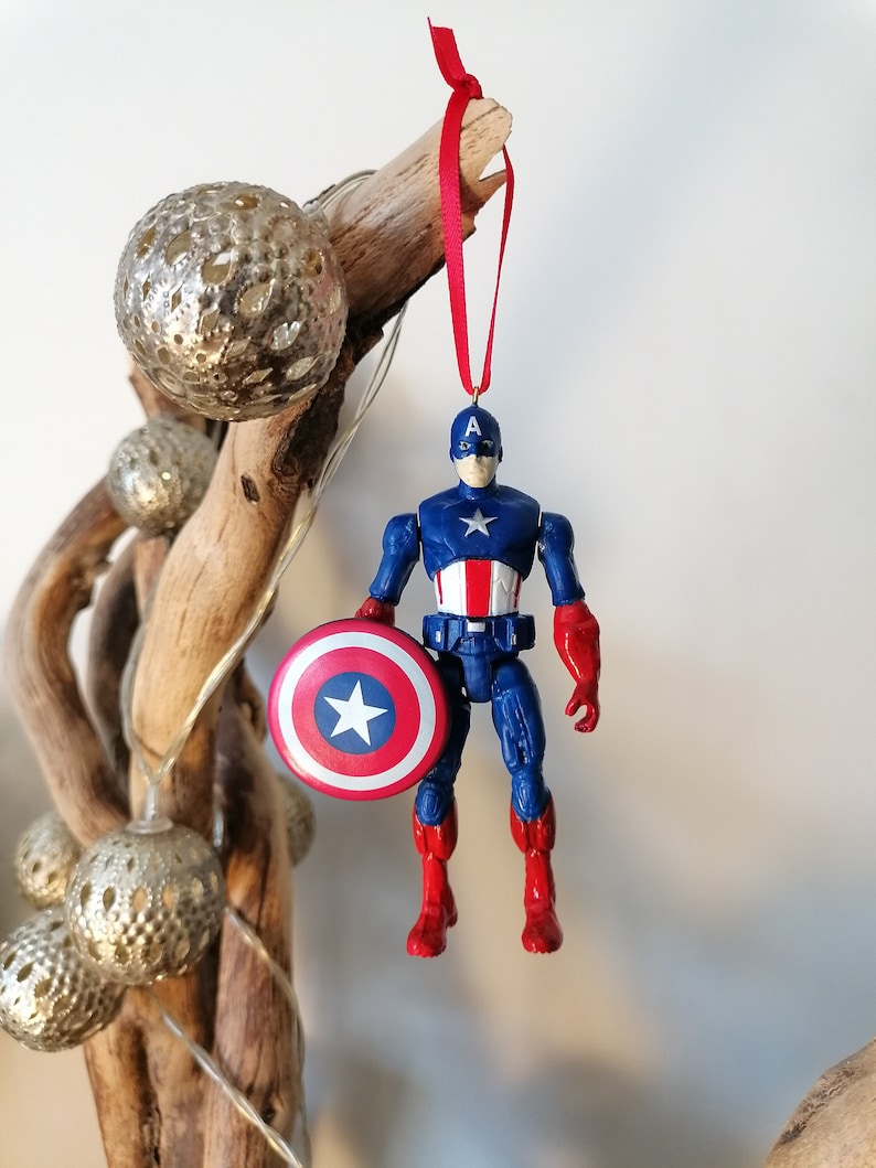 May include: A red, white, and blue Captain America action figure ornament with a red ribbon and a silver star on his shield.