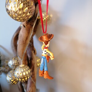 Pu&ograve; includere: Un nastro rosso pende da un ramo con una statuetta di plastica di Woody di Toy Story. Woody indossa il suo cappello da cowboy, il gilet e i jeans caratteristici.