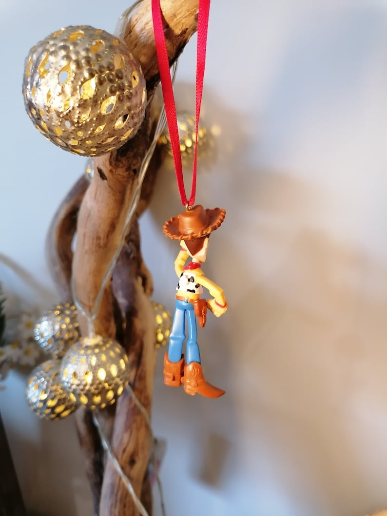 Pu&ograve; includere: Un nastro rosso pende da un ramo con una statuetta di plastica di Woody di Toy Story. Woody indossa il suo cappello da cowboy, il gilet e i jeans caratteristici.