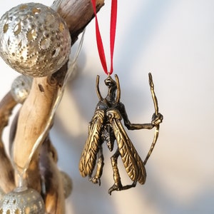May include: A gold-coloured ornament depicting a winged creature with a long, thin staff. The ornament is hanging from a red ribbon.