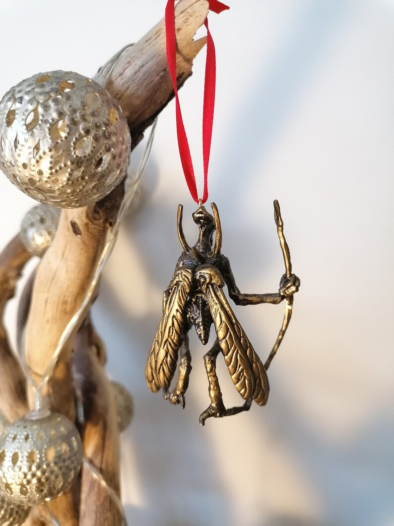 May include: A gold-coloured ornament depicting a winged creature with a long, thin staff. The ornament is hanging from a red ribbon.