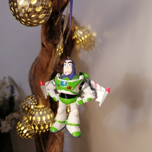 Pu&ograve; includere: Un ornamento di Natale Buzz Lightyear verde e bianco con una tuta da ranger spaziale viola e un'ala di navicella spaziale rossa e bianca. L'ornamento &egrave; appeso a un nastro blu su un ramo di legno.