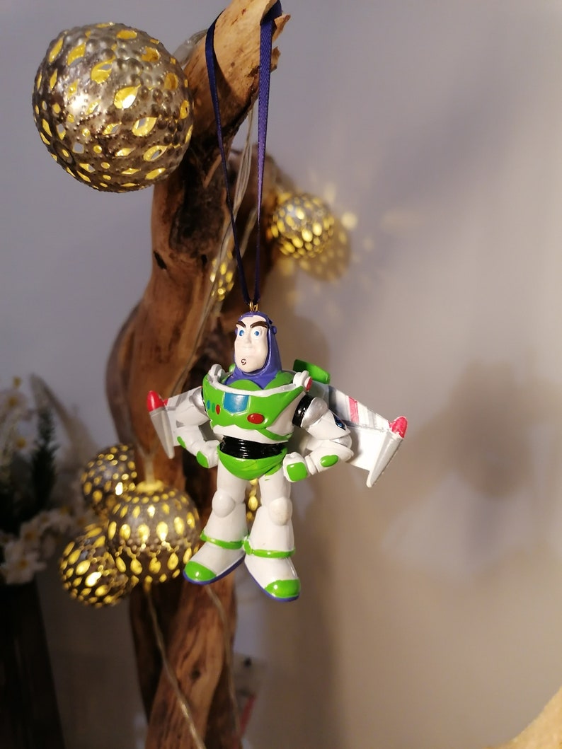 Pu&ograve; includere: Un ornamento di Natale Buzz Lightyear verde e bianco con una tuta da ranger spaziale viola e un'ala di navicella spaziale rossa e bianca. L'ornamento &egrave; appeso a un nastro blu su un ramo di legno.