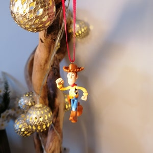 May include: A red string ornament featuring a plastic figurine of Woody from the Toy Story franchise. Woody is wearing his signature cowboy hat, vest, and jeans. He is holding his right arm out in front of him, as if he is running.
