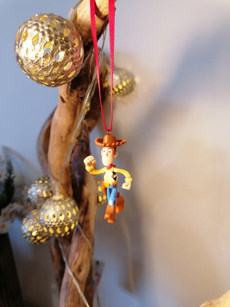 May include: A red string ornament featuring a plastic figurine of Woody from the Toy Story franchise. Woody is wearing his signature cowboy hat, vest, and jeans. He is holding his right arm out in front of him, as if he is running.