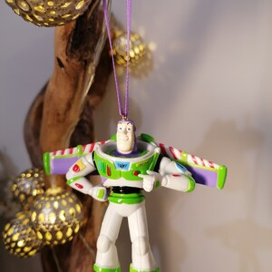 Peut inclure: Ornement de No&euml;l Buzz l'&Eacute;clair, avec une combinaison spatiale blanche et verte et des d&eacute;tails violets. Le soldat jouet est suspendu &agrave; un ruban violet. D&eacute;coration festive.