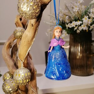Puede incluir: Un adorno de princesa con vestido azul y morado, con una trenza y una cinta azul colgando de una rama con adornos dorados.