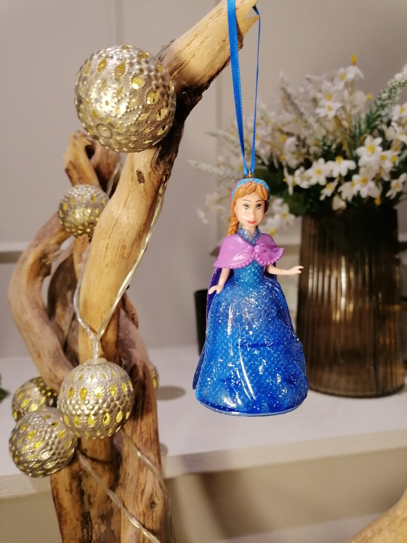 Puede incluir: Un adorno de princesa con vestido azul y morado, con una trenza y una cinta azul colgando de una rama con adornos dorados.