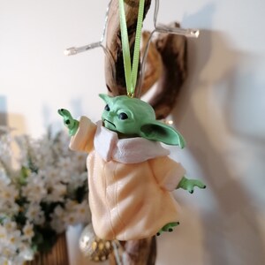 May include: A green and beige Christmas ornament featuring a character from the Star Wars franchise. The ornament is hanging from a green ribbon.