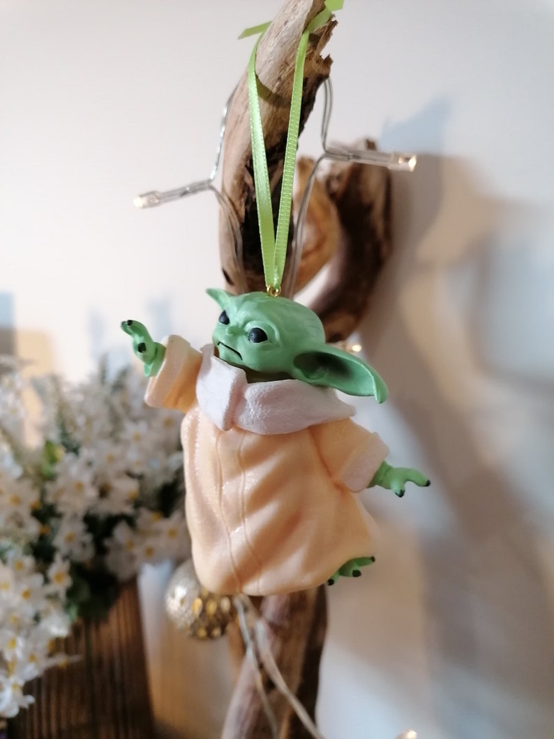 May include: A green and beige Christmas ornament featuring a character from the Star Wars franchise. The ornament is hanging from a green ribbon.
