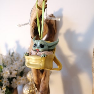 May include: A green and yellow Christmas ornament featuring a character from the Star Wars franchise. The character is sitting in a yellow pouch with a green ribbon attached.