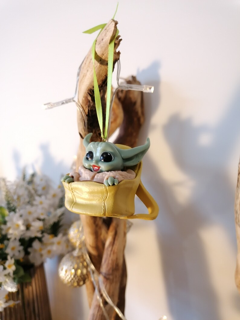 May include: A green and yellow Christmas ornament featuring a character from the Star Wars franchise. The character is sitting in a yellow pouch with a green ribbon attached.