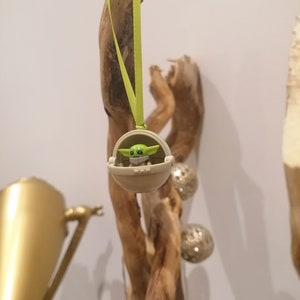 May include: A green and tan Baby Yoda ornament hanging from a light green ribbon. The ornament is in a tan carrier. The background includes a piece of driftwood and silver decorative spheres.
