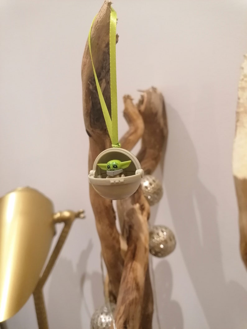 May include: A green and tan Baby Yoda ornament hanging from a light green ribbon. The ornament is in a tan carrier. The background includes a piece of driftwood and silver decorative spheres.