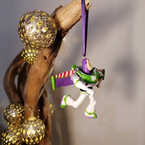 May include: A purple and green plastic toy ornament of Buzz Lightyear from the Toy Story franchise. The ornament is hanging from a purple ribbon on a branch with gold and silver ornaments.