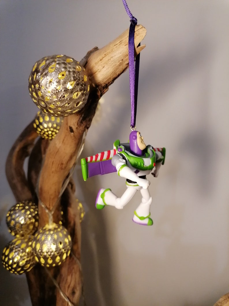 May include: A purple and green plastic toy ornament of Buzz Lightyear from the Toy Story franchise. The ornament is hanging from a purple ribbon on a branch with gold and silver ornaments.