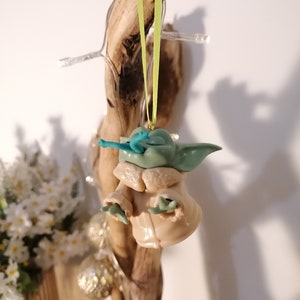 May include: A green and brown Yoda ornament with a blue lightsaber hanging from a green ribbon on a wooden branch.