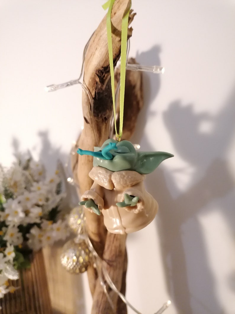 May include: A green and brown Yoda ornament with a blue lightsaber hanging from a green ribbon on a wooden branch.