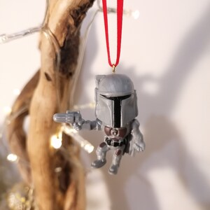 May include: A grey and black Star Wars Mandalorian Christmas ornament with a red ribbon hanging from a branch.