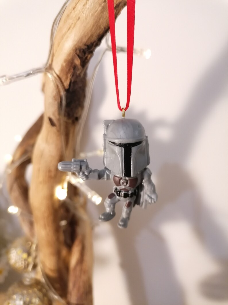 May include: A grey and black Star Wars Mandalorian Christmas ornament with a red ribbon hanging from a branch.