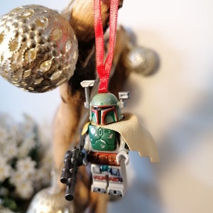 May include: A green and brown Lego Star Wars Boba Fett minifigure ornament with a tan cape hanging from a red ribbon.