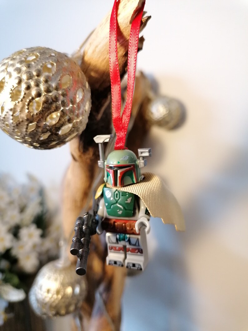May include: A green and brown Lego Star Wars Boba Fett minifigure ornament with a tan cape hanging from a red ribbon.