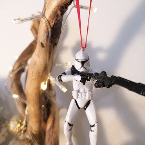 Puede incluir: Un adorno navide&ntilde;o de Star Wars Stormtrooper blanco y negro colgado de una cinta roja en una rama con luces.