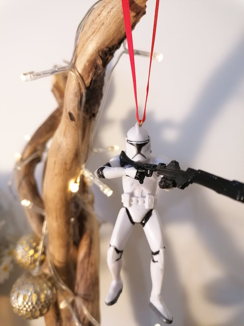 Puede incluir: Un adorno navide&ntilde;o de Star Wars Stormtrooper blanco y negro colgado de una cinta roja en una rama con luces.
