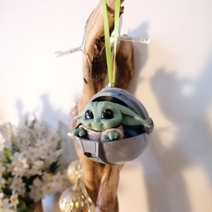 May include: A green and gray Christmas ornament featuring a character from the Star Wars franchise, The Mandalorian. The character is sitting in a pod-like structure with a green ribbon attached.