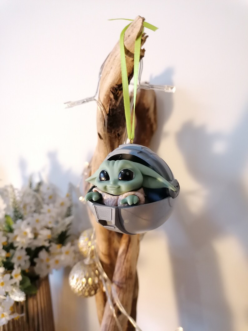 May include: A green and gray Christmas ornament featuring a character from the Star Wars franchise, The Mandalorian. The character is sitting in a pod-like structure with a green ribbon attached.