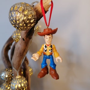 May include: A red and brown plastic toy figurine of Woody from Toy Story hanging from a red ribbon. Woody is wearing his signature cowboy hat, vest, and jeans.