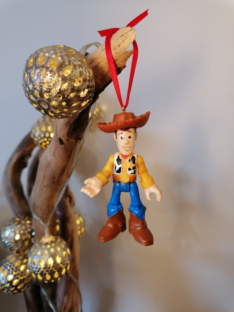 May include: A red and brown plastic toy figurine of Woody from Toy Story hanging from a red ribbon. Woody is wearing his signature cowboy hat, vest, and jeans.
