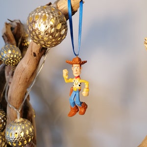 May include: A blue ribbon hangs from a branch with a plastic figurine of Woody from Toy Story. Woody is wearing his signature cowboy hat, vest, and jeans. He is holding his arm out and his legs are bent as if he is running.