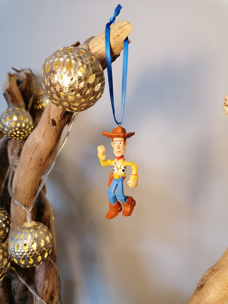 May include: A blue ribbon hangs from a branch with a plastic figurine of Woody from Toy Story. Woody is wearing his signature cowboy hat, vest, and jeans. He is holding his arm out and his legs are bent as if he is running.